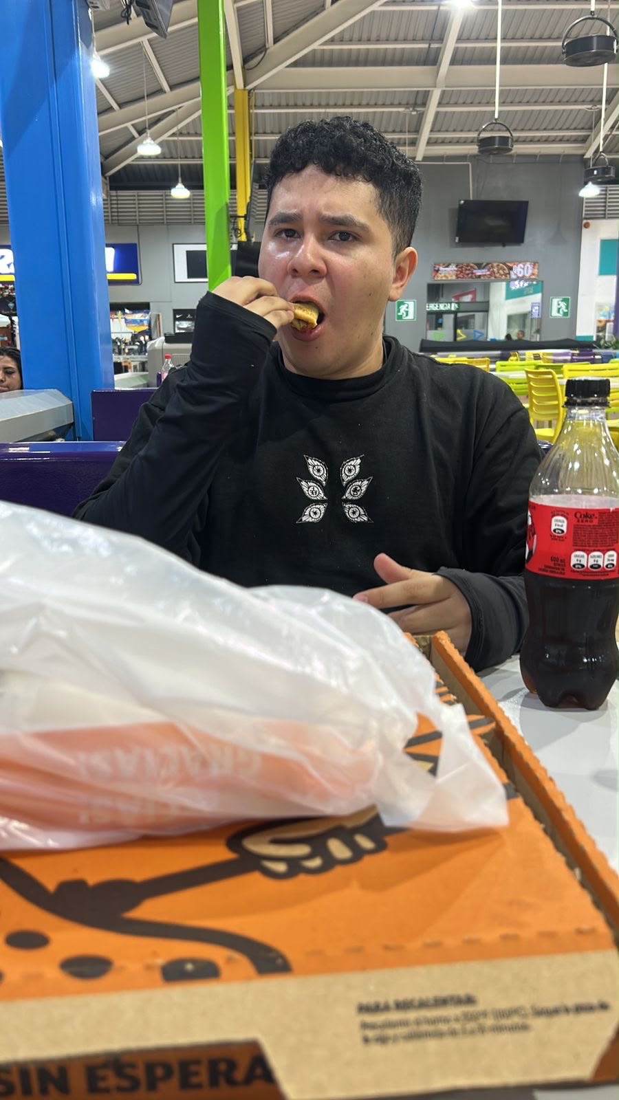 El beneficiario dando una mordida en un food court, con una caja de pizza y una Coca-Cola en la mesa