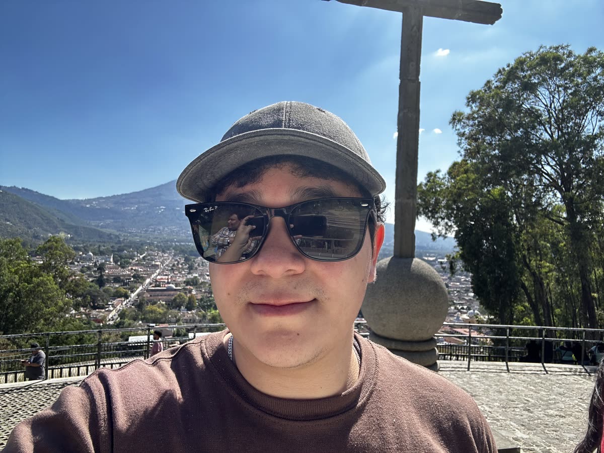 Selfie del beneficiario con gorra y lentes oscuros en un mirador, con árboles, una cruz de piedra y un volcán de fondo