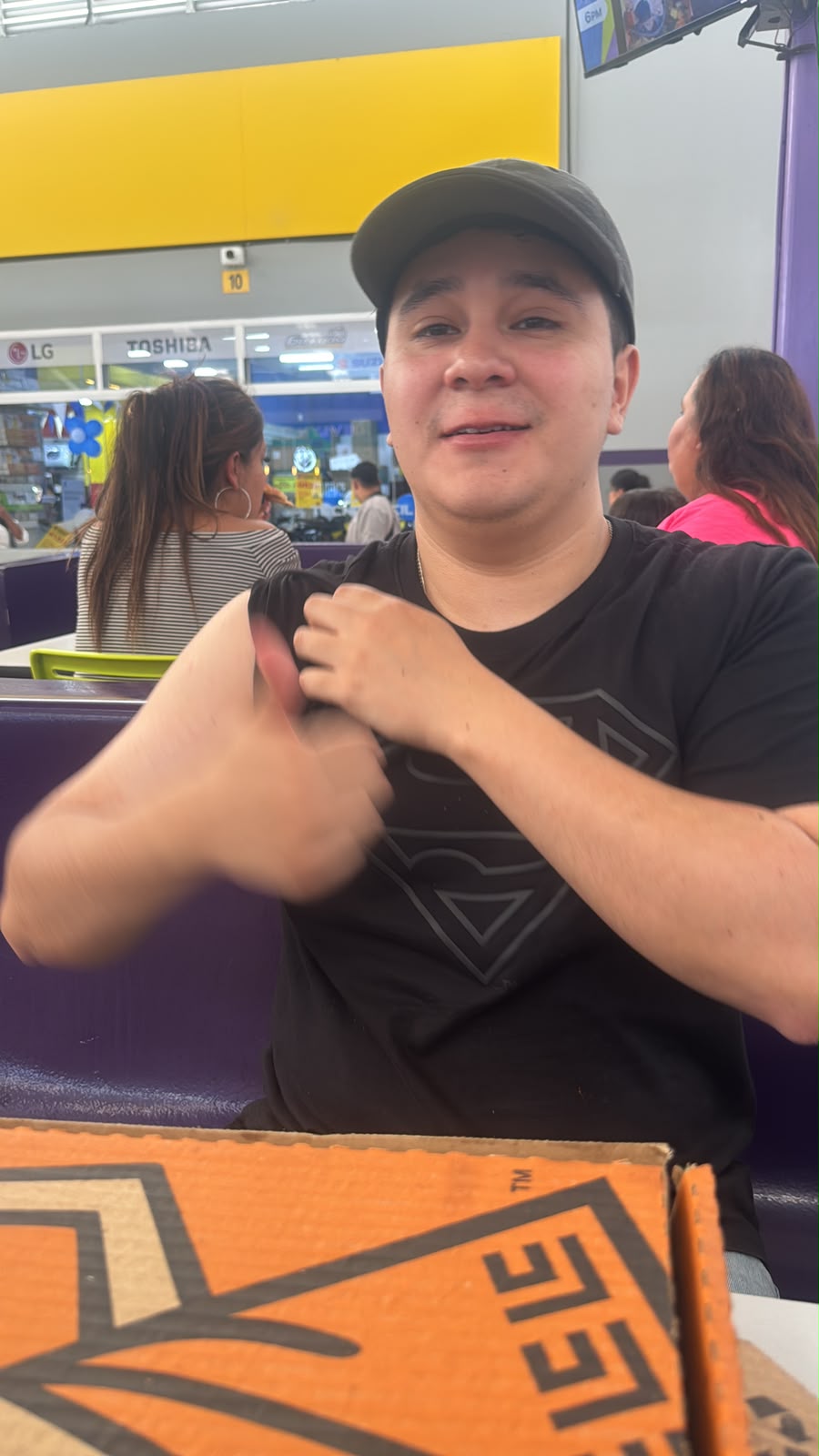 El beneficiario en un food court, con gorra, haciendo pulgar arriba junto a una caja de pizza